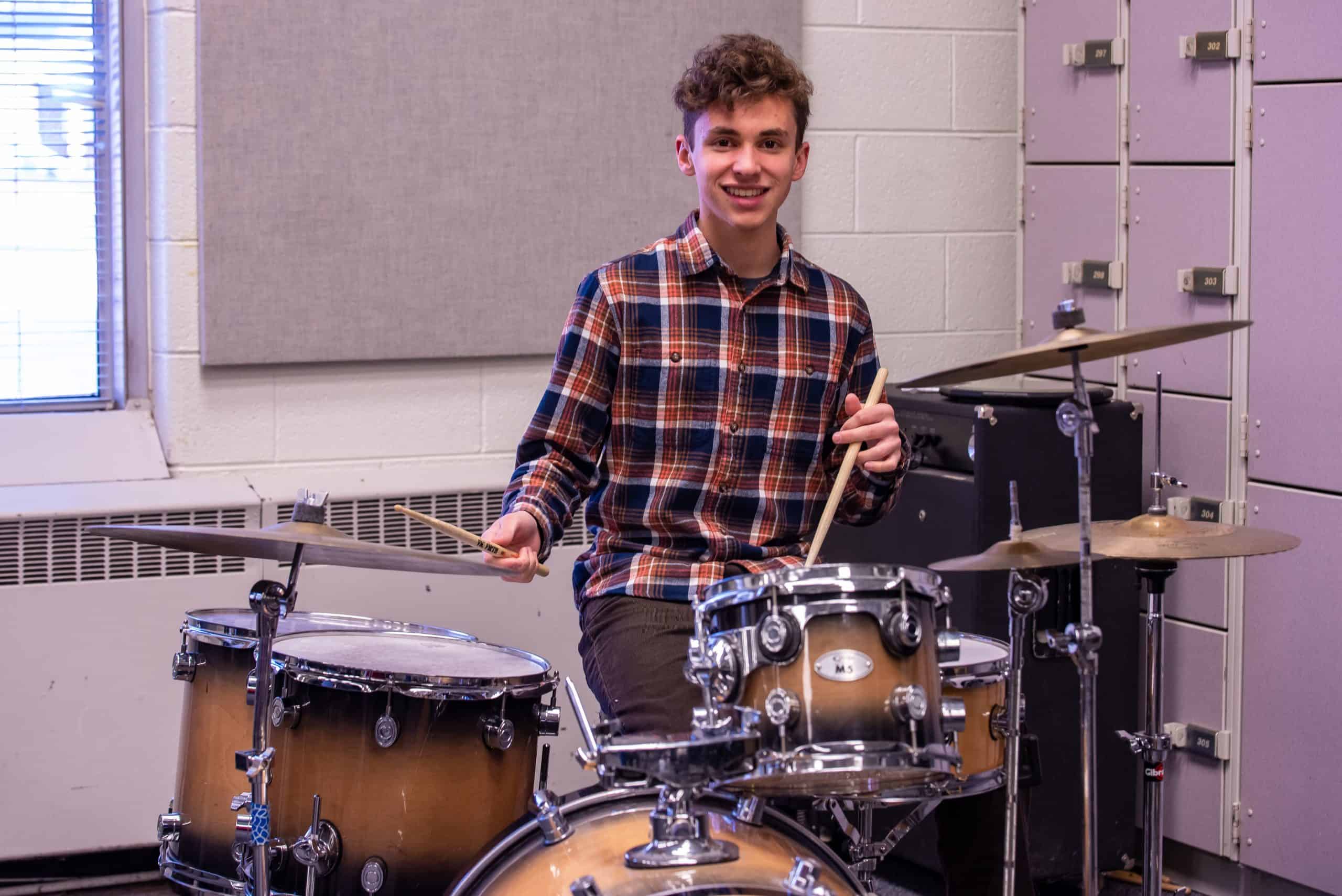 High school male, Simon Von Hatten, sitting at drumset holding drumsticks in each hand and smiling at the camera.