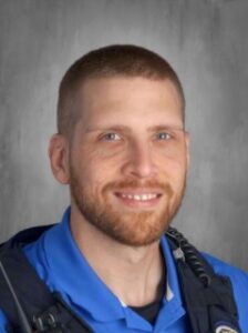 Smiling male officer with short hair and a beard, wearing a blue uniform, against a gray background.