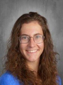 A smiling individual with curly hair and glasses, wearing a blue shirt, against a gray background.