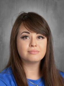 A young woman with long brown hair and winged eyeliner, wearing a blue shirt, against a gray background.
