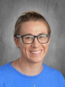 Smiling person with short hair wearing glasses and a blue shirt, against a gray background.
