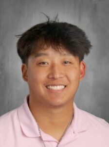 A young man with short, dark hair smiles while wearing a pink shirt against a neutral background.