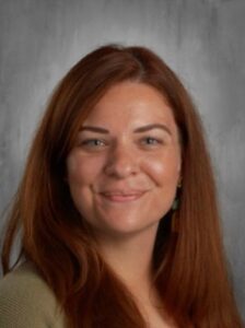 A woman with long, wavy red hair smiles at the camera, wearing green earrings and a light sweater.