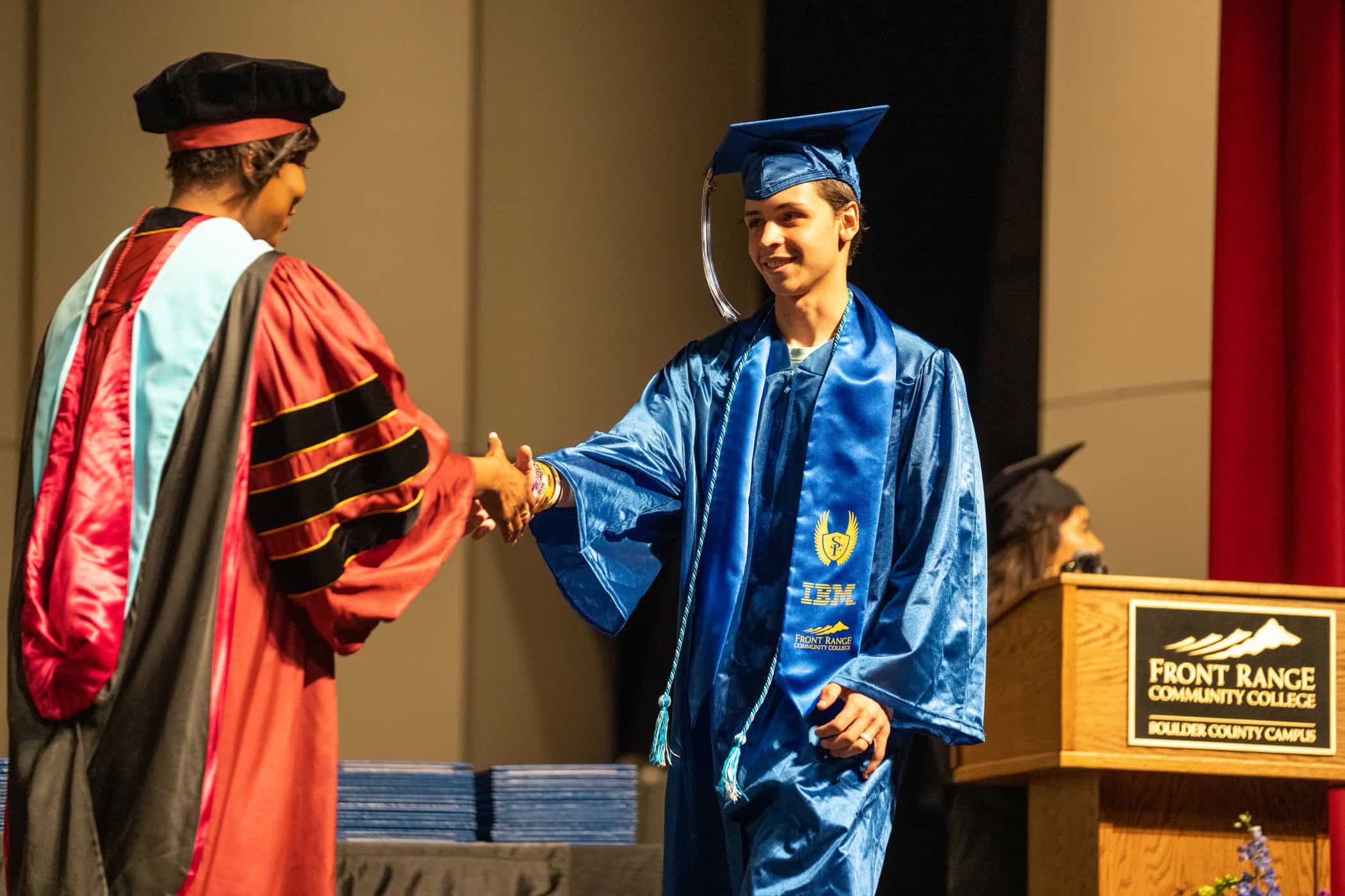 A graduate in a blue gown shakes hands with a woman in academic regalia during a graduation ceremony.