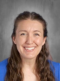 Smiling woman with long brown hair wearing a blue top, set against a gray background.