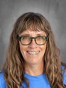 A smiling woman with long, wavy hair and glasses, wearing a blue shirt, against a gray background.