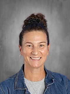 A smiling woman with a bun hairstyle, wearing a denim jacket and gray shirt, set against a gray background.