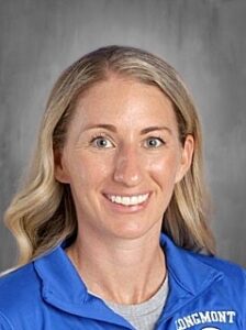 Smiling woman with long blonde hair wearing a blue sports jacket, set against a gray background.