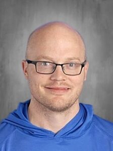 A smiling bald man wearing glasses and a blue hoodie, against a gray background.