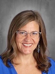 Smiling woman with glasses and earrings, wearing a blue top, set against a gray background.