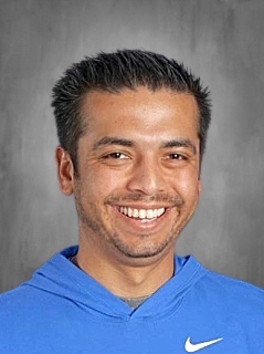 Smiling man with short dark hair wearing a blue hoodie against a gray background.