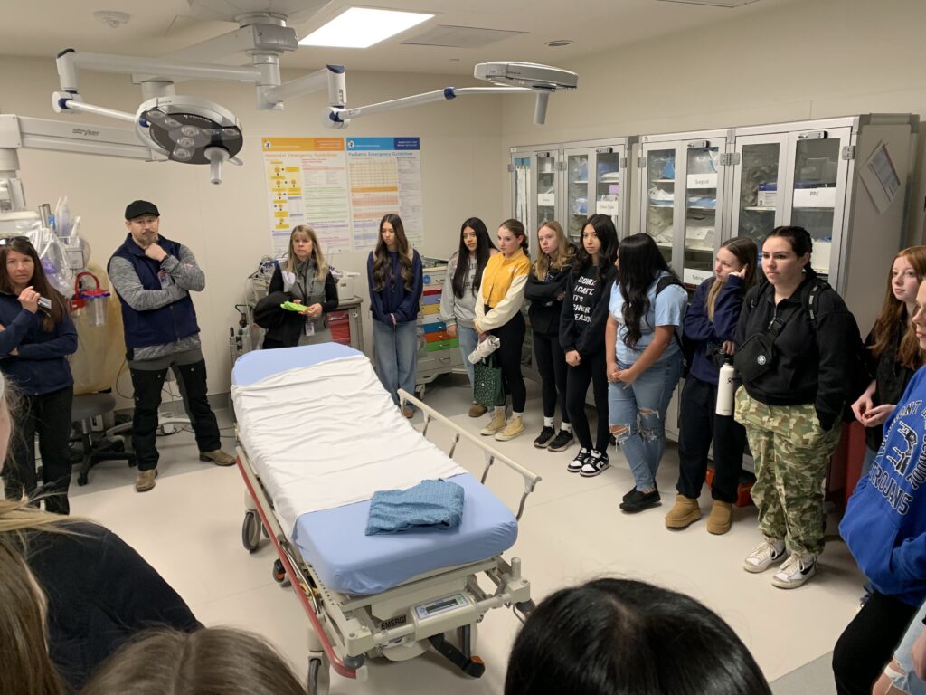 A group of students observes a demonstration in a hospital room with a medical bed and equipment.