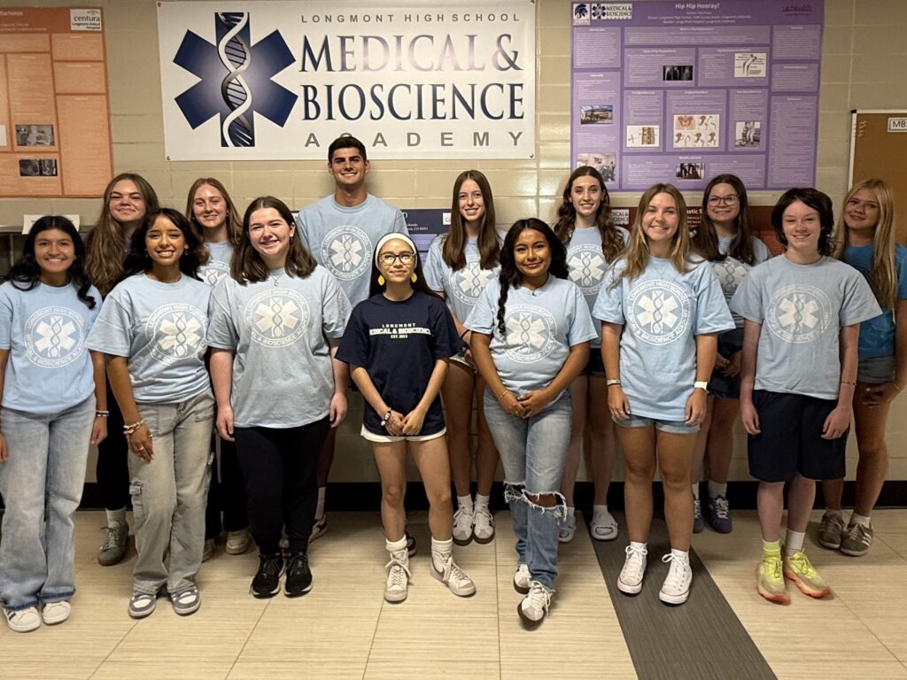 A group of students stands in front of a Medical & Bioscience Academy sign, all wearing matching light blue shirts.