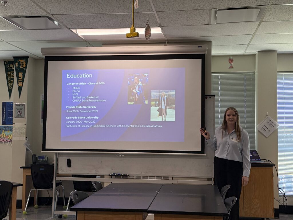 A person stands beside a presentation screen detailing their education background at a classroom setting.