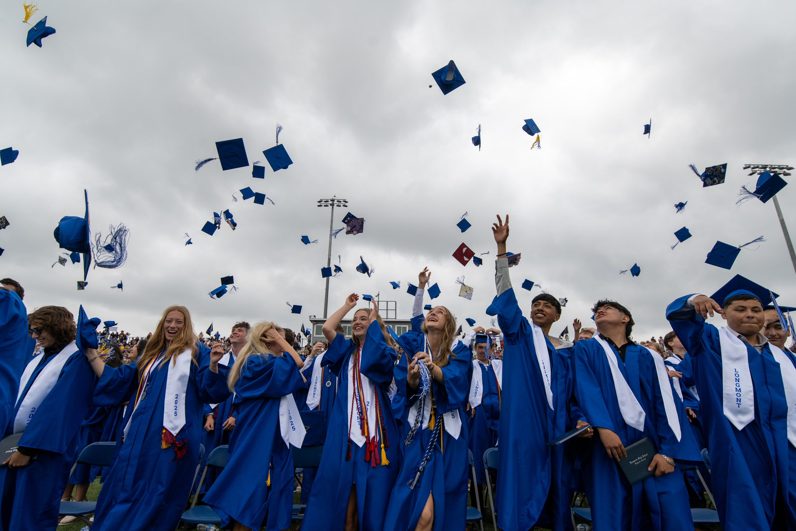 Graduates in blue gowns joyfully toss their caps into the air during a graduation ceremony under cloudy skies.
